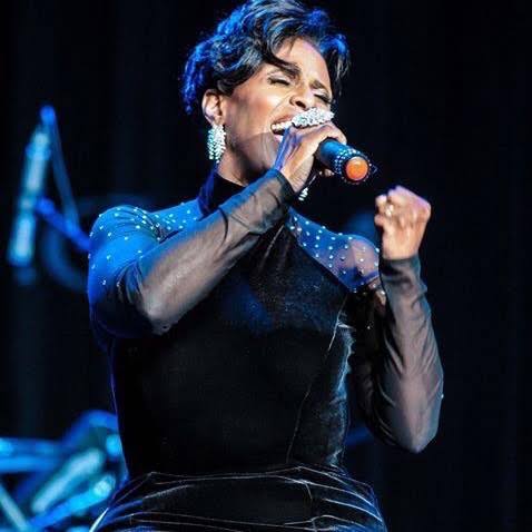 Yolanda Rabun performing live, eyes closed mid-phrase, singing into a handheld microphone in a dark studded-sleeve top